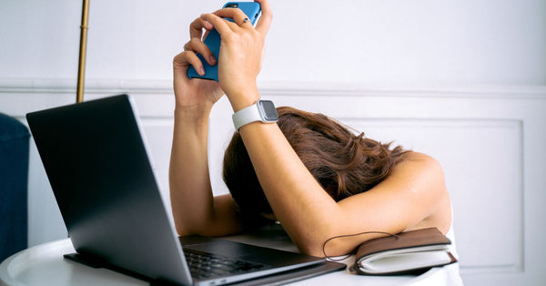 Tired woman resting head on desk next to laptop holding phone showing digital detox tips for adults